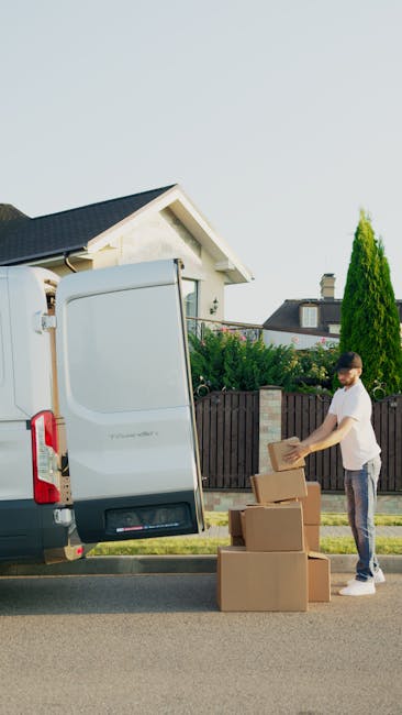 A man wearing a blue beanie hat, a navy blue and red waterproof jacket, black trousers, and white sneakers with black and red accents is sitting on the edge of an open van door during a home relocatio