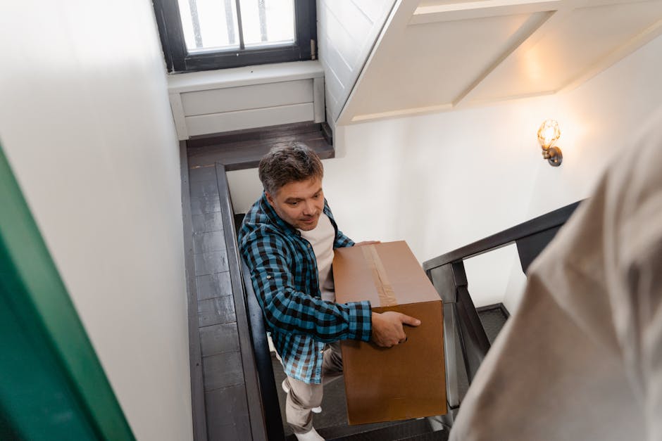 A man with short brown hair, wearing a blue and black checked shirt over a white t-shirt, is carrying a cardboard box during a home relocation. He is positioned on a staircase inside a property, with the staircase railing visible in the foreground. The stairs are dark-colored, possibly wooden, and lead down from an upper floor near a window that allows natural light into the space. Inside the property, the walls are white, and there is a small wall-mounted light fixture on the right side, emitting warm illumination. The box he is holding appears to be sealed with packing tape. The environment suggests an organized packing and moving process, with [COMPANY_NAME] providing professional removals services, supporting efficient furniture transport and load handling during the house move between floors, as shown in this stair carry scenario for the Streatham Common flat removals page.