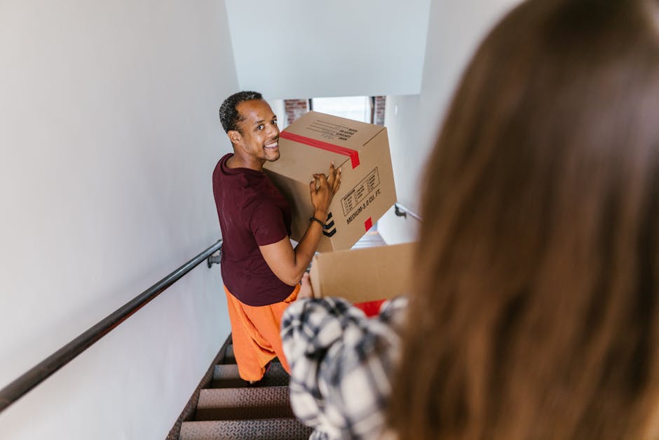 A man wearing a maroon t-shirt and orange trousers is fishing a medium-sized cardboard box with red tape from inside a residential property. He is smiling and looking upward as he carries the box down a staircase, which has a black metal handrail on the left side. The staircase is carpeted with a brown patterned runner, and the wall on the left is painted white, while the wall behind him shows a red brick window sill with natural light coming through a window. In the foreground, the back of a woman with long brown hair, wearing a plaid shirt, is visible as she hands another cardboard box to the man, indicating a home relocation process associated with house removals. The scene captures the logistics of packing and furniture transport, typical of a professional removals service like Man With a Van Streatham, during a stair carry solution operation for a flat or house move.