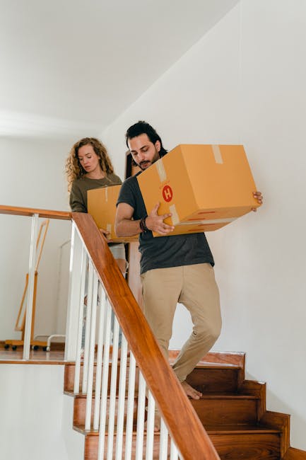 A man with short brown hair, wearing a blue and black checked shirt over a white t-shirt, is carrying a cardboard box during a home relocation. He is positioned on a staircase inside a property, with the staircase railing visible in the foreground. The stairs are dark-colored, possibly wooden, and lead down from an upper floor near a window that allows natural light into the space. Inside the property, the walls are white, and there is a small wall-mounted light fixture on the right side, emitting warm illumination. The box he is holding appears to be sealed with packing tape. The environment suggests an organized packing and moving process, with [COMPANY_NAME] providing professional removals services, supporting efficient furniture transport and load handling during the house move between floors, as shown in this stair carry scenario for the Streatham Common flat removals page.