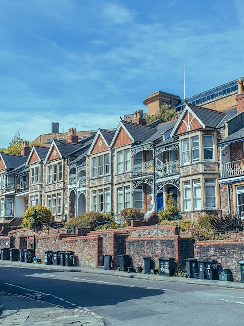 A row of Victorian-style terraced houses on Streatham High Road, featuring ornate metalwork balconies, bay windows, and gabled roofs with brick and white detailing. The houses are set behind a brick wall, with small landscaped front gardens and shrubbery. Several black wheelie bins are lined along the pavement in front of the properties. The sky above is clear and blue, with some wispy clouds. The scene suggests a residential area where home relocations and furniture transport might take place, with the intact street environment supporting house removals services such as those offered by Man With a Van Streatham.