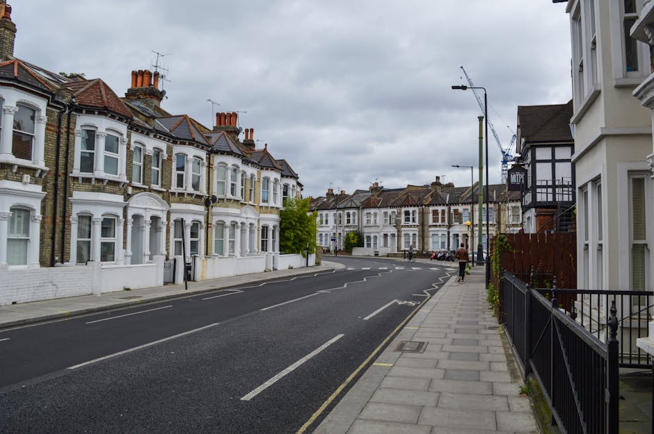A street scene in Streatham with a row of Victorian-style terraced houses on the left, featuring white and brick facades, bay windows, and red tiled roofs. The pavement on the right side is lined with metal railings and residential porches. A man in dark clothing is walking along the sidewalk, carrying a small box or bag. The road is empty of vehicles, with visible road markings indicating parking restrictions, and a few cars are parked further down. Overhead, on the right, there are tall streetlights and a construction crane visible in the distance against a cloudy sky, suggesting ongoing development in the area. This scene reflects a typical residential area suitable for house removals and furniture transport, with clear access to homes for moving services like those offered by Man With a Van Streatham.