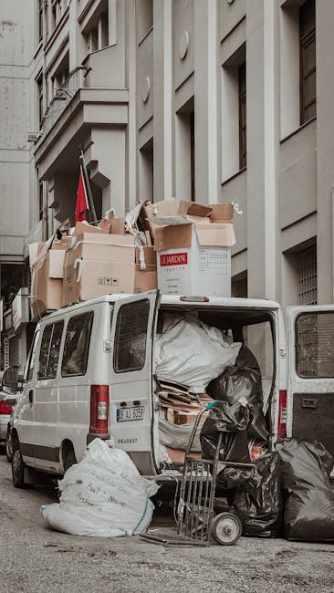 The back of a white commercial van parked on a street in a residential area, with its rear doors open showing an interior filled with various packed cardboard boxes, black plastic trash bags, and roll